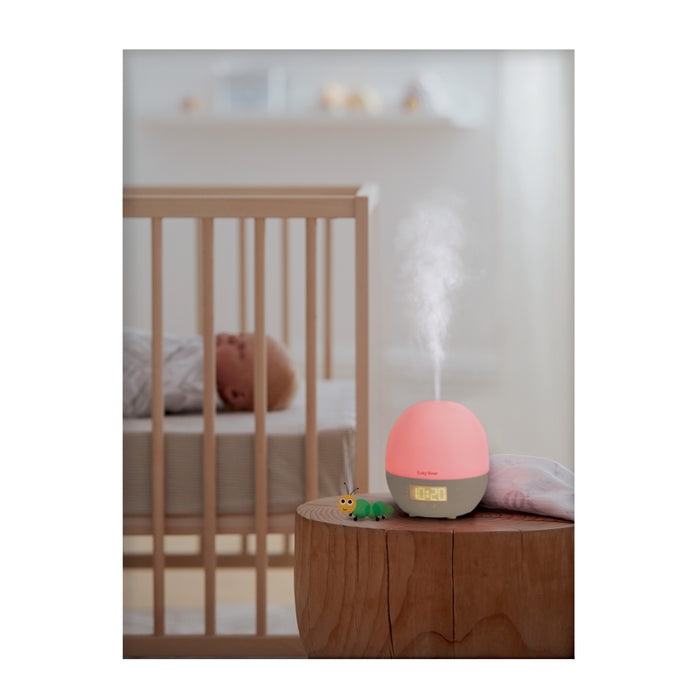 Euky Bear Sweet Dreams Baby Sleep Aid in pink on a bedside table, releasing mist, with digital clock display glowing, next to a sleeping baby in a crib.